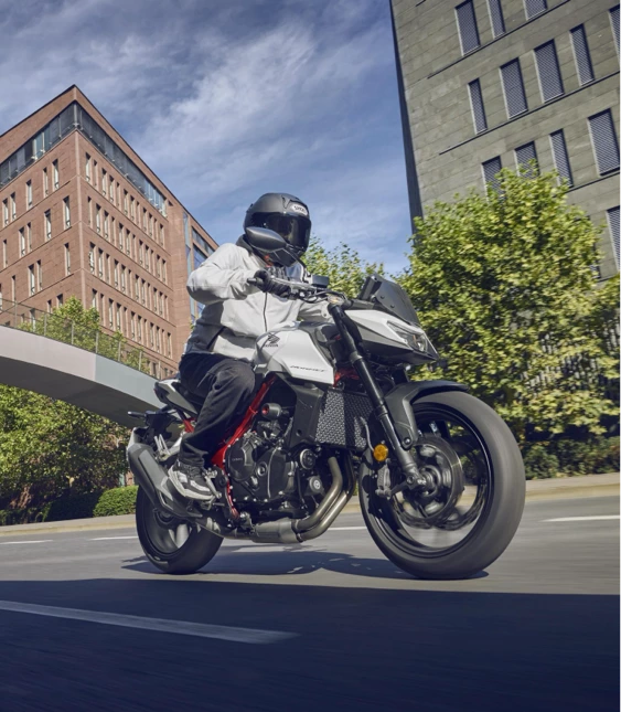 Motorcyklist kör på väg i stadsmiljö med grönskande träd i bakgrunden.