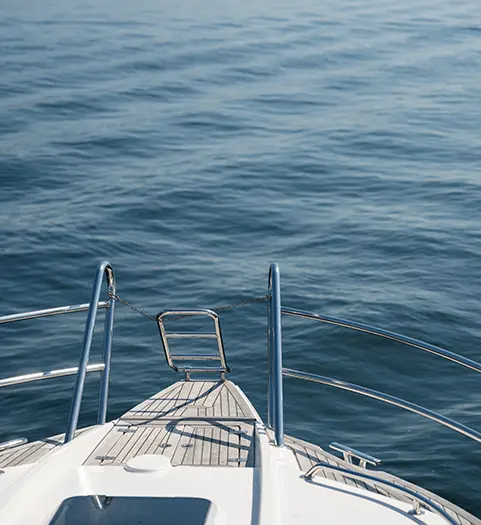 Bild på fören av en båt. Trädäck och badstege samt relingar syns i bild mot havet.