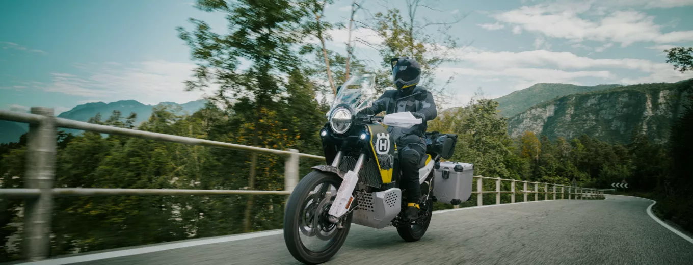 Motorcyklist i full skyddsutrustning kör på kurvig landsväg med berg och grönskande natur i bakgrunden.