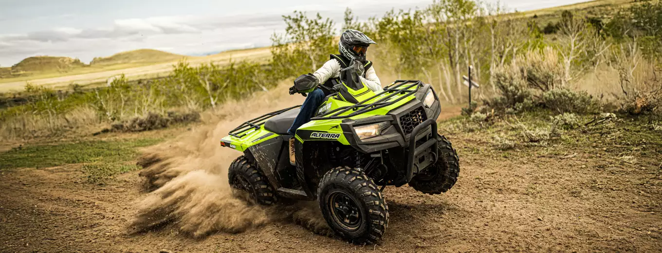 Person kör grön ATV från Arctic Cat i terräng med dammoln bakom.