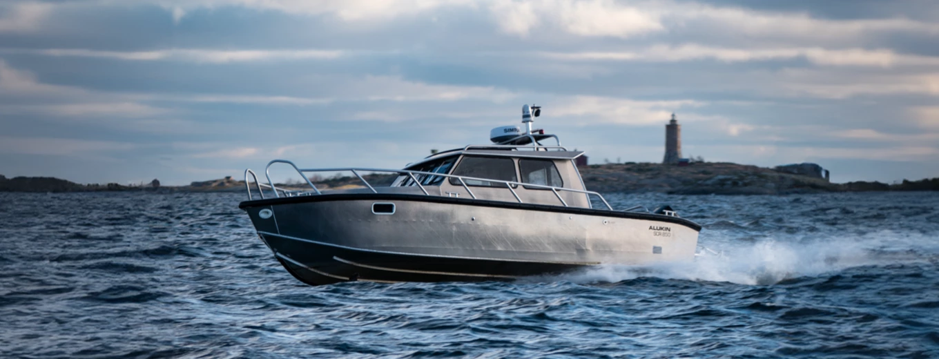 Aluminiumbåt av märket Alukin med kabin kör snabbt över ett vågigt hav med en fyr i bakgrunden.