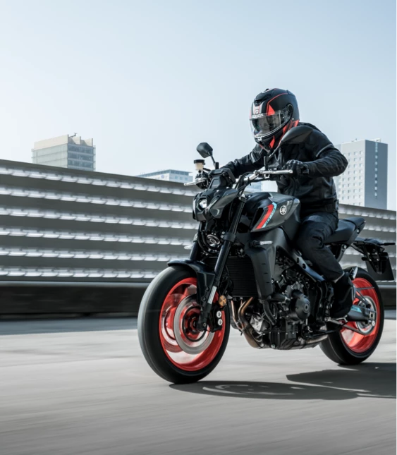 En motorcyklist i full svart skyddsutrustning kör en svart motorcykel med röda detaljer på en upphöjd väg med stadssilhuetten i bakgrunden