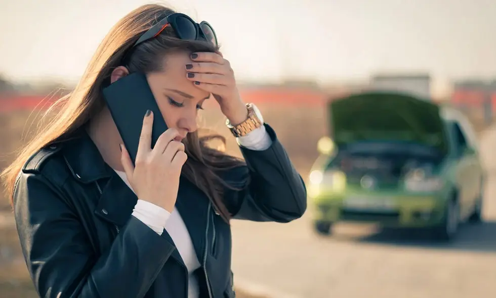 En kvinna som fått motorstopp pratar i telefon med en öppen motorhuv i bakgrunden