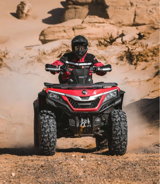 Förare i hjälm kör röd ATV genom sandig terräng, med damm som yr runt fordonet i ett öppet landskap.