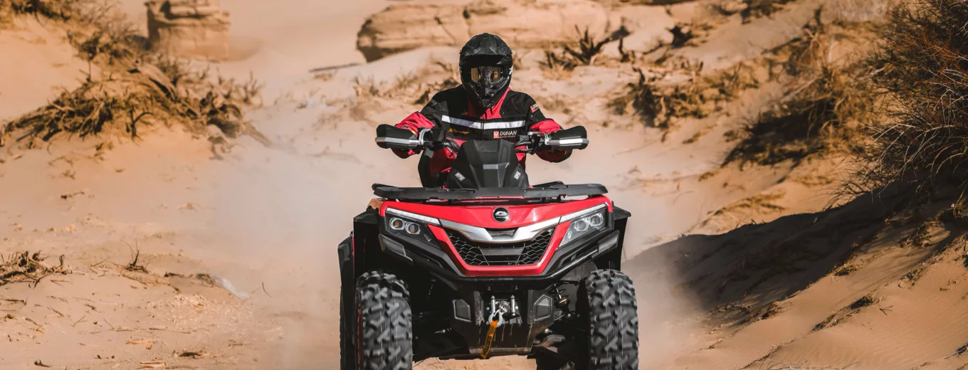 Förare i hjälm kör röd ATV genom sandig terräng, med damm som yr runt fordonet i ett öppet landskap.