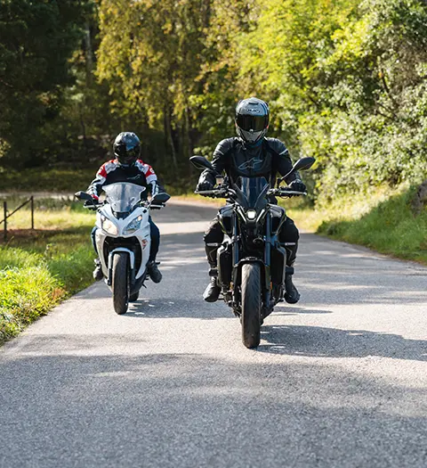 Två motorcyklister i full körutrustning på en landsväg omgiven av grönska, med den främre föraren på en svart motorcykel följd av en förare på en vit och röd sportmotorcykel
