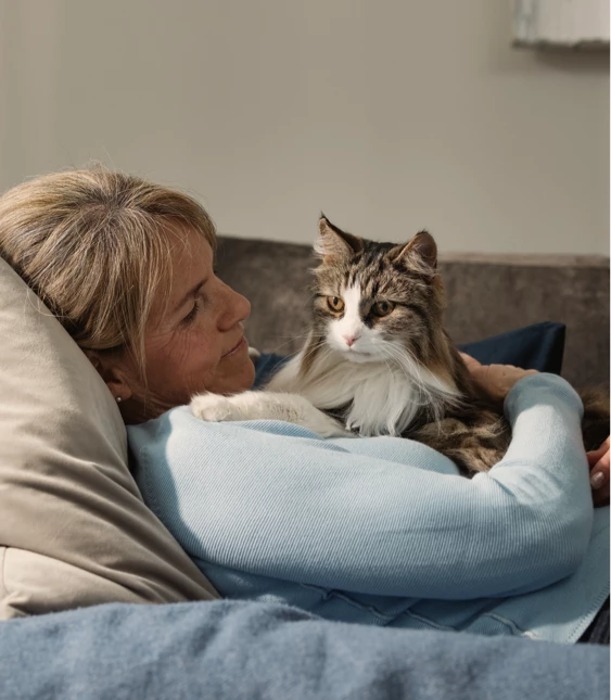 En kvinna ligger tillbakalutad på en soffa och håller om en fluffig katt, båda tittar mot kameran med avslappnade uttryck.