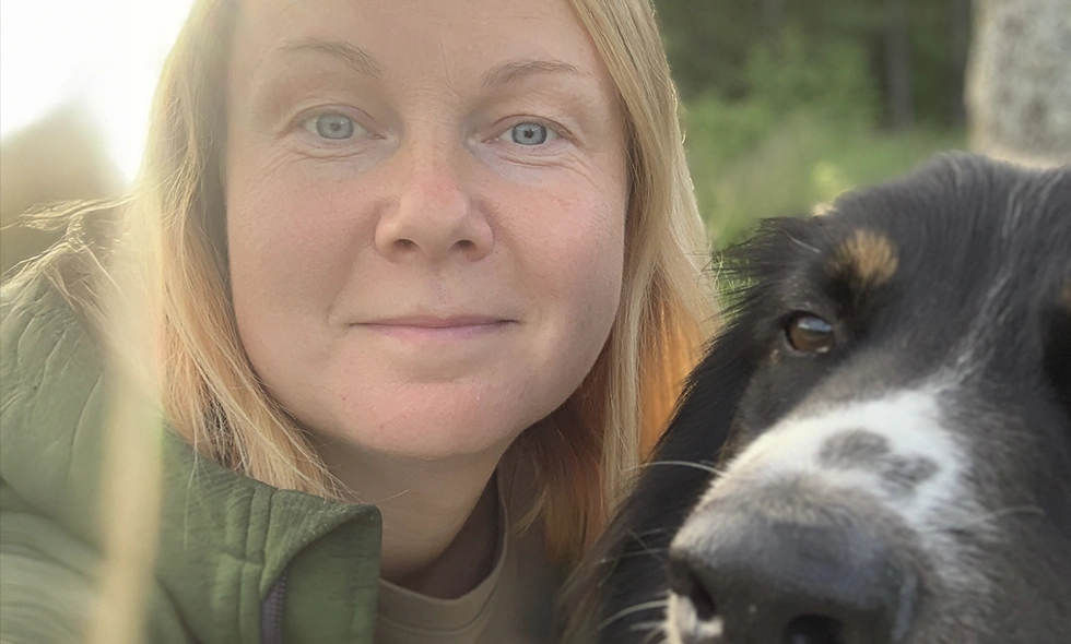 Närbild på en kvinna med blont hår i grön jacka som tar en selfie tillsammans med en svart och vit hund utomhus i naturen.