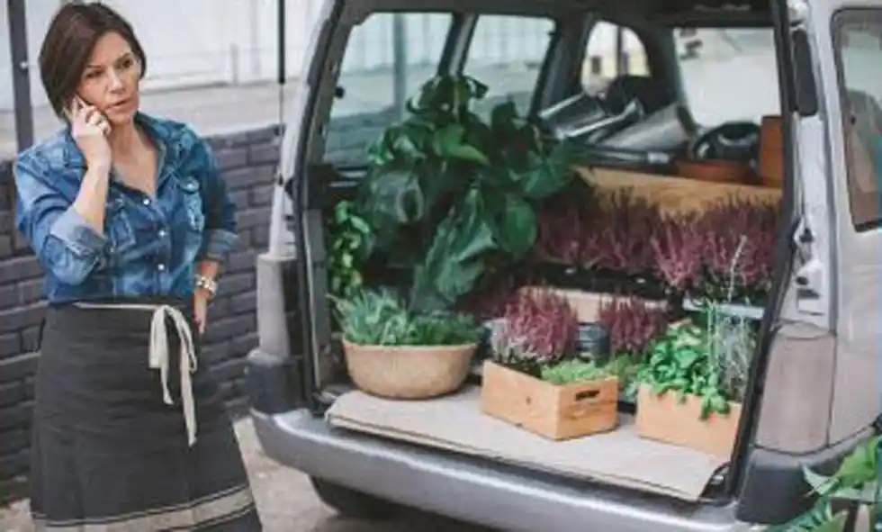 En kvinna stå och pratar i mobilen bakom en minivan fylld med växter.