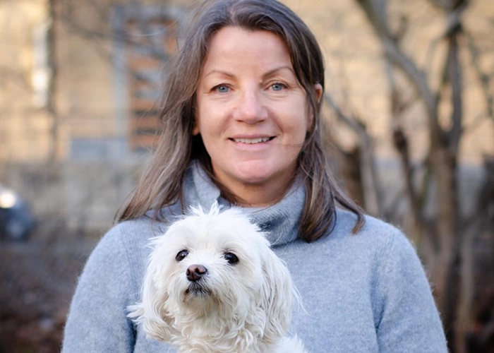 Kvinna som ler in i kameran med vit hund i knät.)