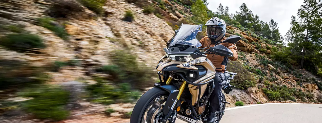 Motorcyklist kör en äventyrsmotorcykel på en slingrande bergsväg med klippor och grönska i bakgrunden.