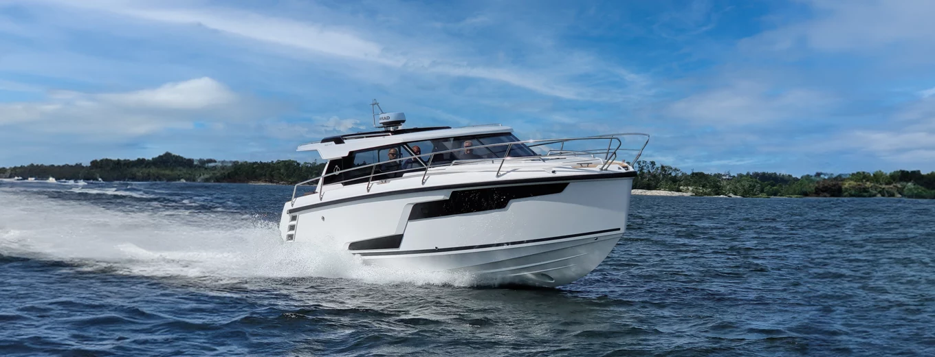 Motorbåt av märket Aquador färdas snabbt över vattnet nära en kustlinje med träd i bakgrunden.