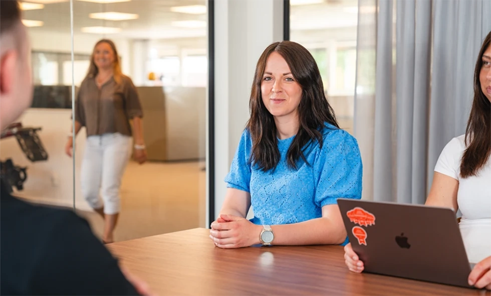 Tre personer sitter vid ett mötesbord i kontorsmiljö, en fjärde person närmar sig i bakgrunden.