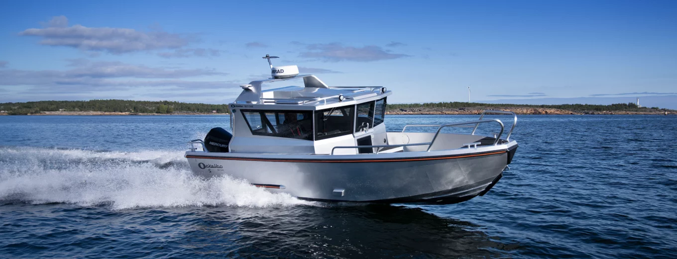 Aluminiumbåt av märket Ockelbo med kabin kör snabbt över en sjö med kustlinje och skog i bakgrunden.