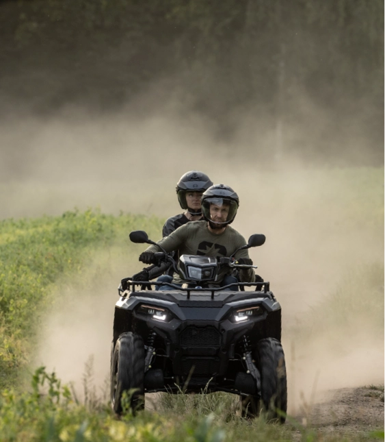 Två personer sitter på en ATV som rör sig i riktning mot kameran med grönt gräs i bakgrunden.