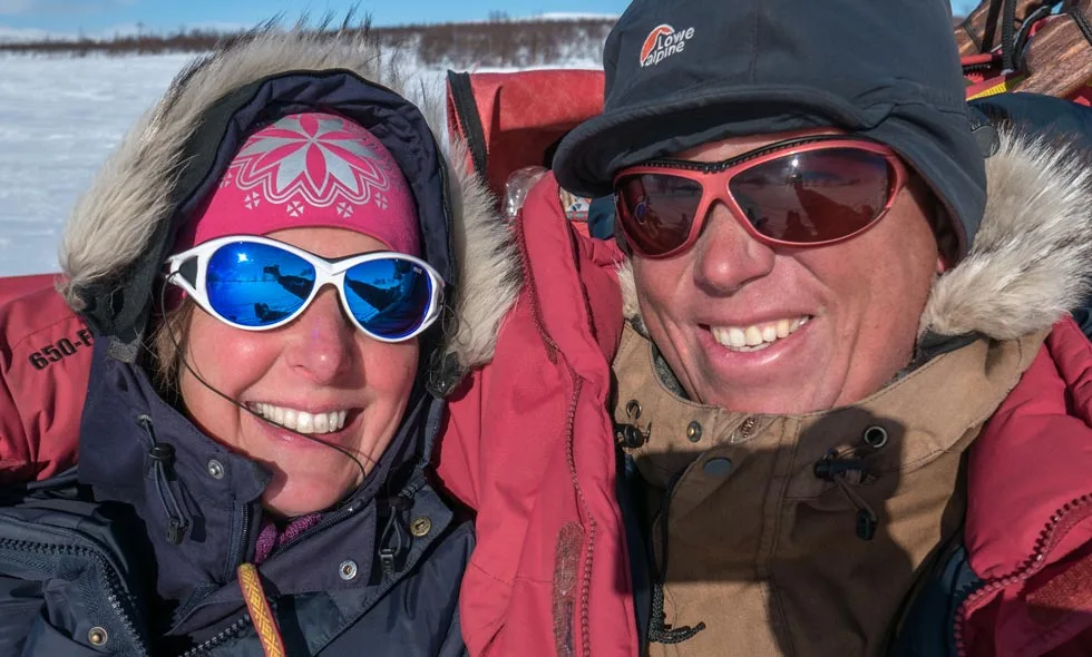 Ett par i vinterkläder med solglasögon och mössor ler mot kameran i ett kallt, snöigt landskap)
