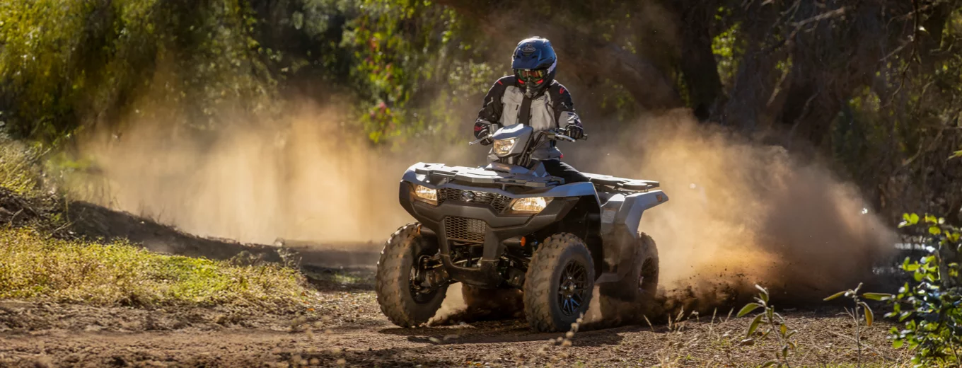 Förare på grå ATV från Suzuki kör på dammig skogsväg.