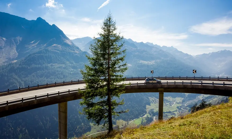  Bil på bergsvägsbro med bergskedja i bakgrunden och en ensam tall framför