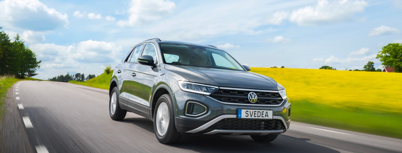 En modern grå Volkswagen med registreringsskylten "SVEDEA" kör längs en landsväg med lummig grönska och ett gult fält i bakgrunden under en klarblå himmel