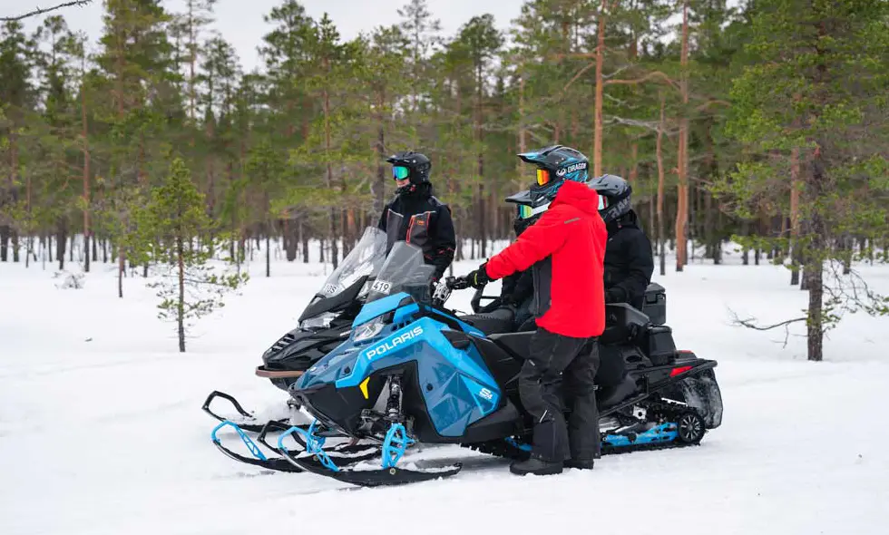 Fyra personer med hjälmar står vid två snöskotrar i ett snötäckt skogslandskap, redo för en tur.