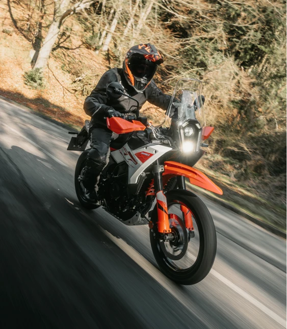 Motorcyklist i svart hjälm kör orange äventyrsmotorcykel genom kurva på skogsväg.