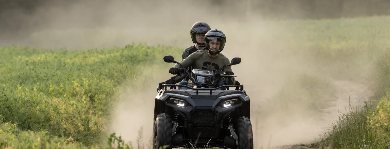 Två personer sitter på en ATV som rör sig i riktning mot kameran med grönt gräs i bakgrunden.