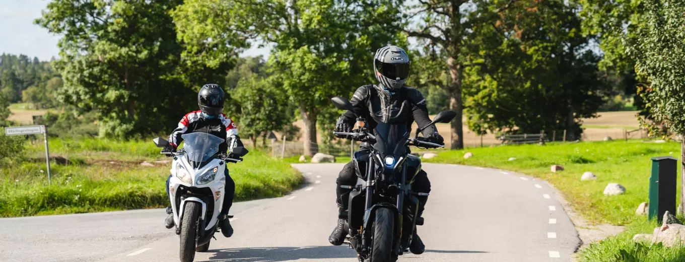 Två motorcyklister, en på en vit sportmotorcykel och en på en svart cruiser, kör på en landsväg omgiven av grönskande natur och fält