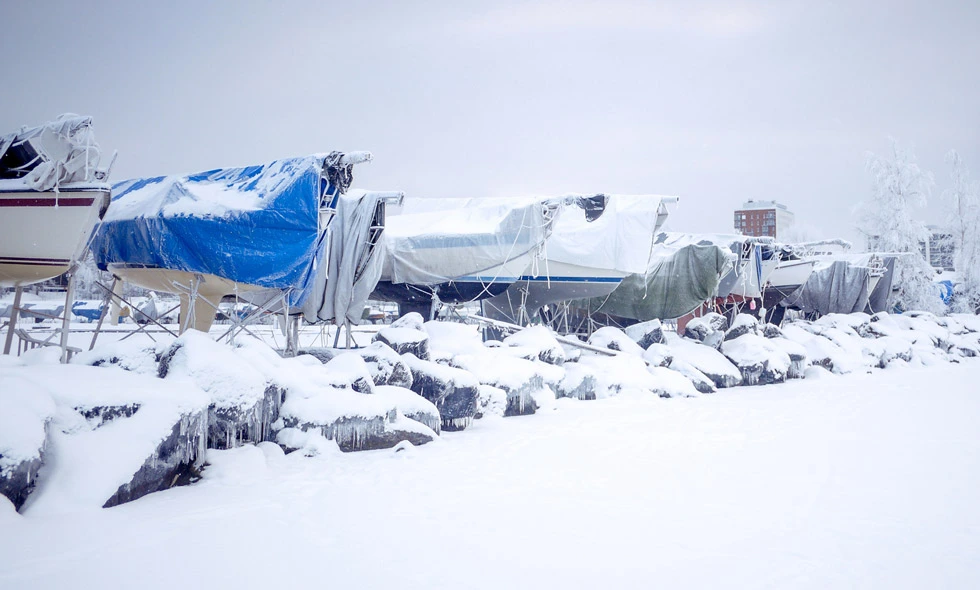 Båtar täckta med snö och presenningar i ett vinterlandskap, byggnad i bakgrunden