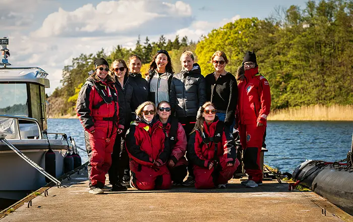 Ett gruppfoto av elva kvinnor, några i röda flytoveraller, stående och sittande på en brygga vid vattnet en solig dag.)
