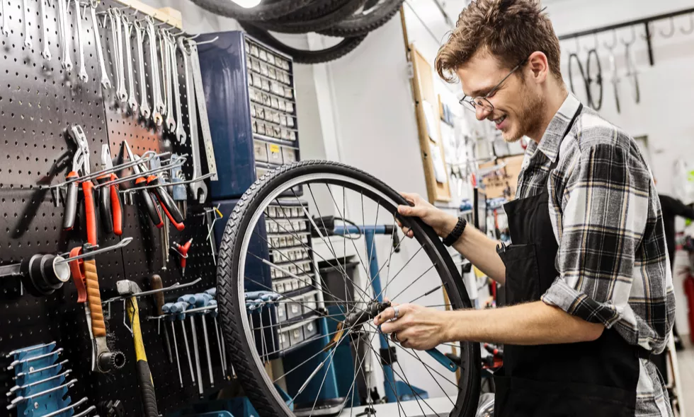 Leende cykelmekaniker justerar ett hjul i verkstad med vägg full av verktyg i bakgrunden.