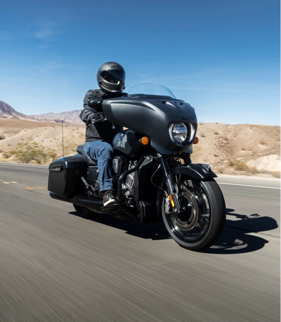 Motorcyklist kör på öde landsväg genom ökenlandskap med berg i bakgrunden under klarblå himmel.