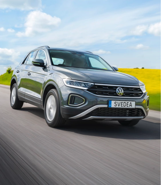 En modern grå Volkswagen med registreringsskylten "SVEDEA" kör längs en landsväg med lummig grönska och ett gult fält i bakgrunden under en klarblå himmel
