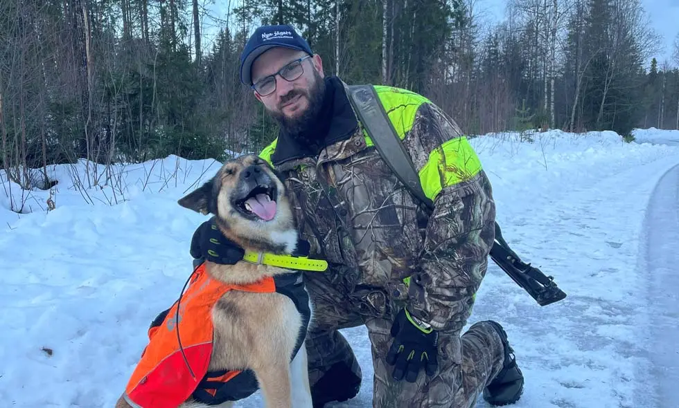 Man i jaktutrustning knäböjer bredvid en hund med väst