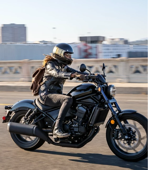 En förare på en svart cruiser-motorcykel kör över en bro i stadsmiljö, med urban skyline i bakgrunden