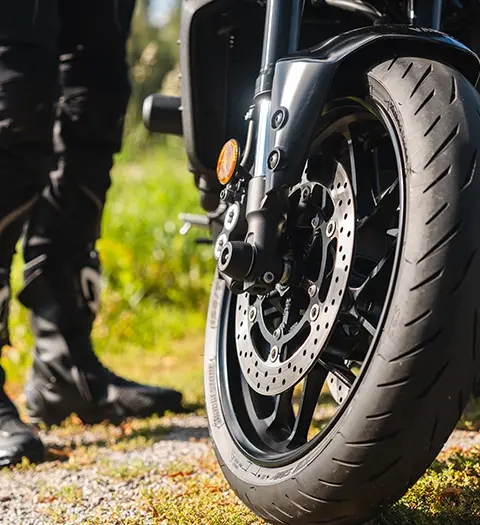 Framhjulet på en parkerad motorcykel, fokus på skivbromsen och däcket, med en person i motorcykelkläder som står i bakgrunden