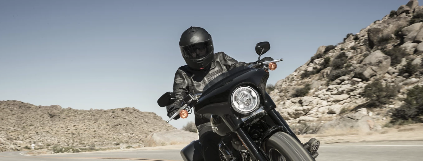 En motorcyklist i svart utrustning kör en svart roadster-motorcykel på en ökenväg, med branta klippor i bakgrunden