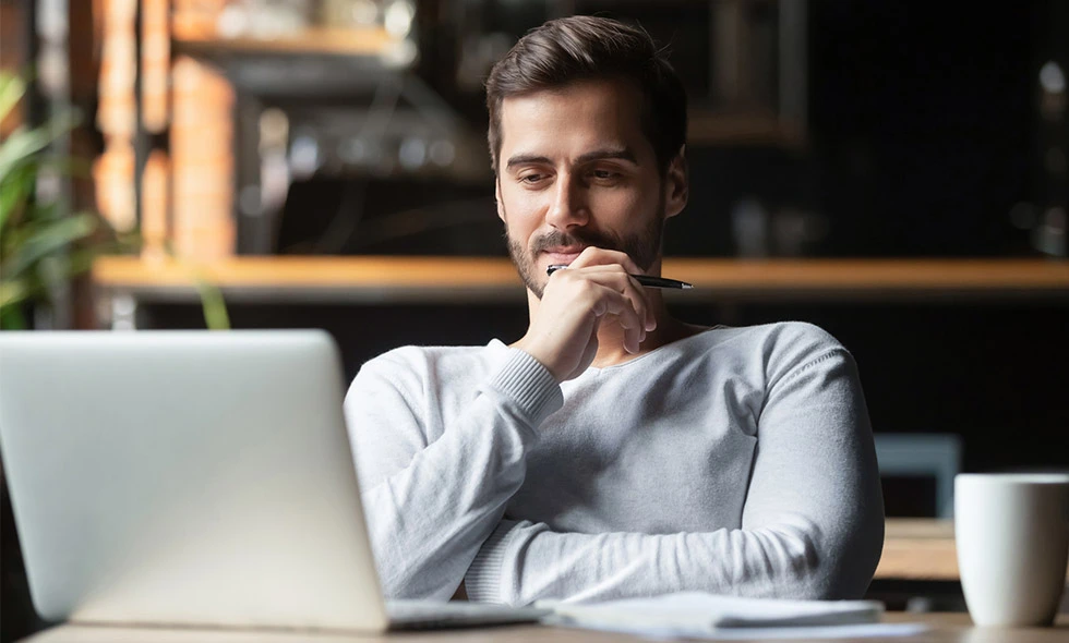 En man i grå tröja funderar koncentrerat framför sin laptop på ett kafé