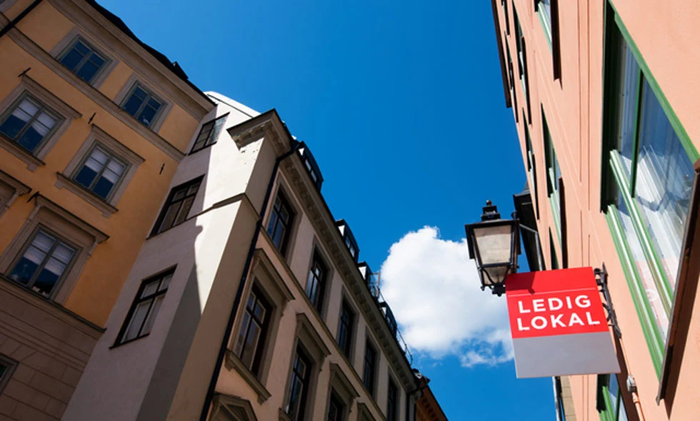 En röd skylt med texten 'LEDIG LOKAL' hänger på en svart lyktstolpe framför färgglada byggnader under en klarblå himmel