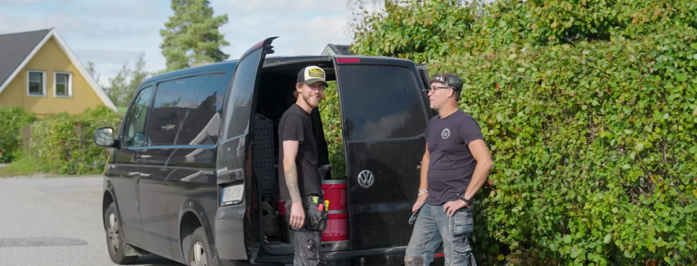 Två män står och samtalar bredvid en öppen svart minivan vid en landsväg, med grönska och ett gult hus i bakgrunden.
