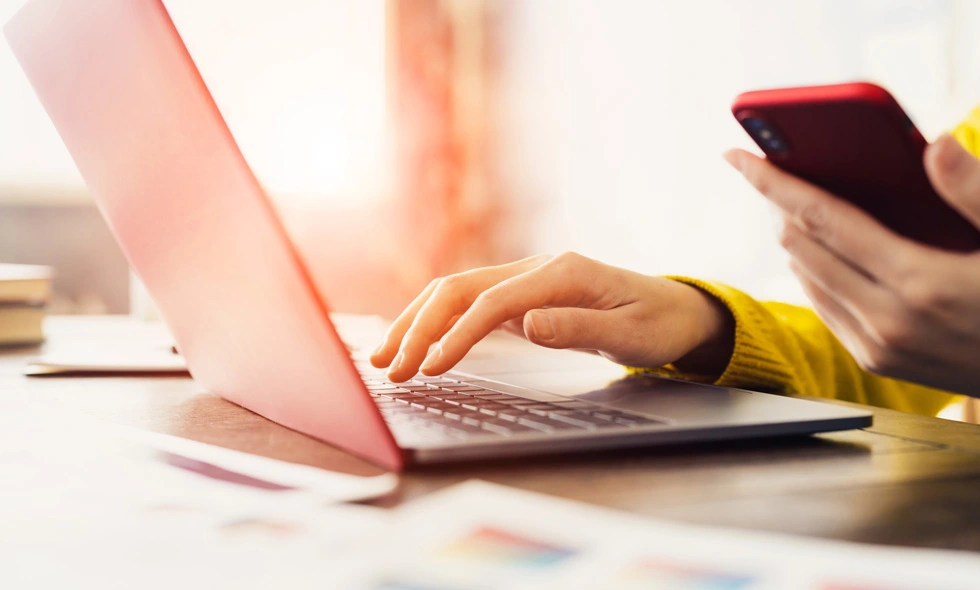 En person som sitter vid ett bord med en laptop och en smartphone i händerna, med ett fönster som släpper in naturligt ljus i bakgrunden