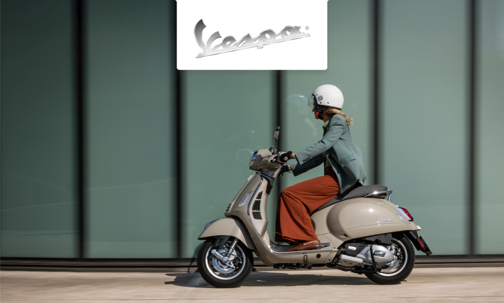 Person kör beige Vespa-scooter längs modern byggnad i stadsmiljö.