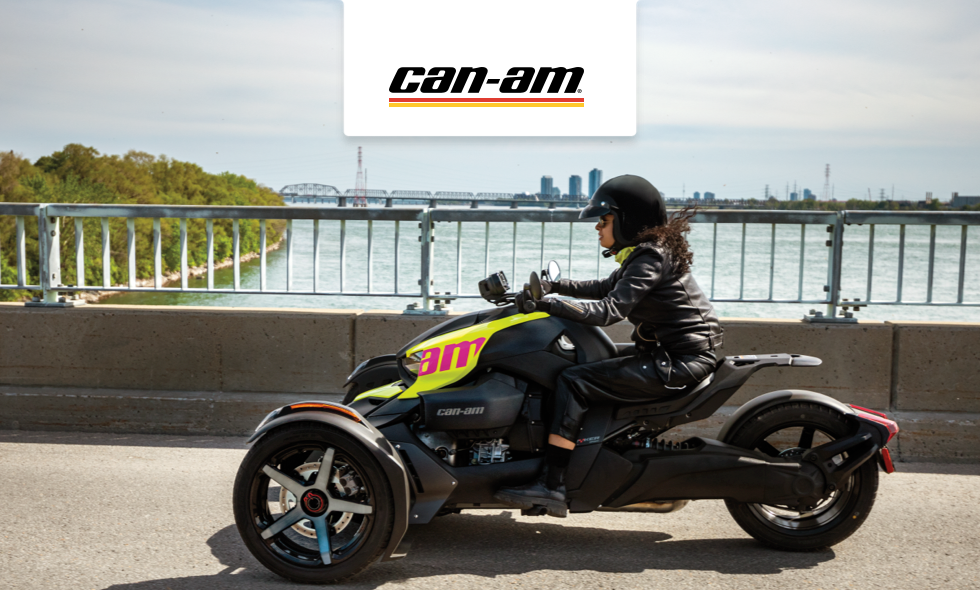 Person kör en Can-Am trehjulig motorcykel över bro med stad och vatten i bakgrunden.