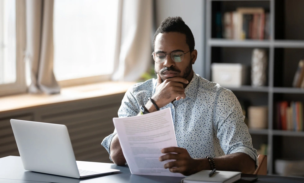 En man i glasögon granskar noggrant ett dokument medan han sitter vid sitt skrivbord med en laptop framför sig