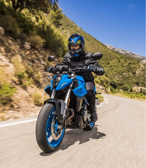 Motorcyklist kör på en solig landsväg med berg och naturlandskap i bakgrunden.