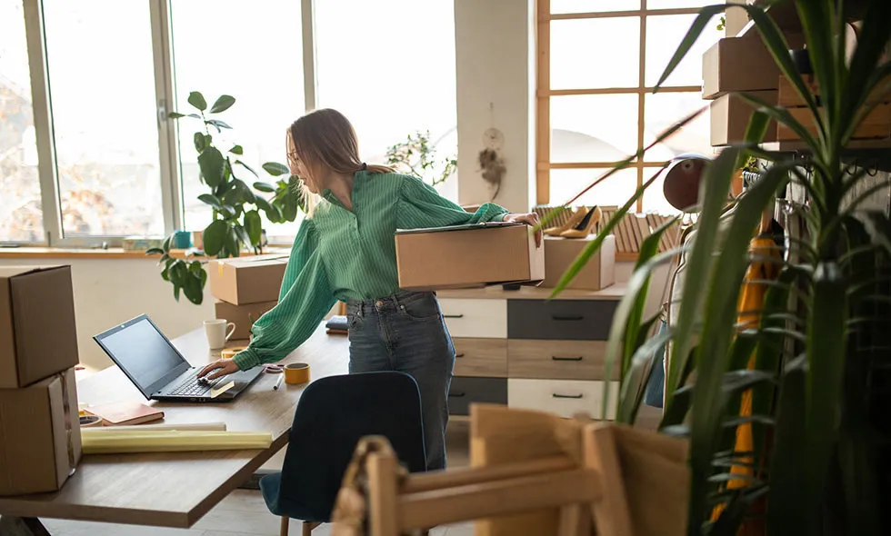 Kvinna packar lådor framför en dator på sitt soliga kontor med många gröna växter.