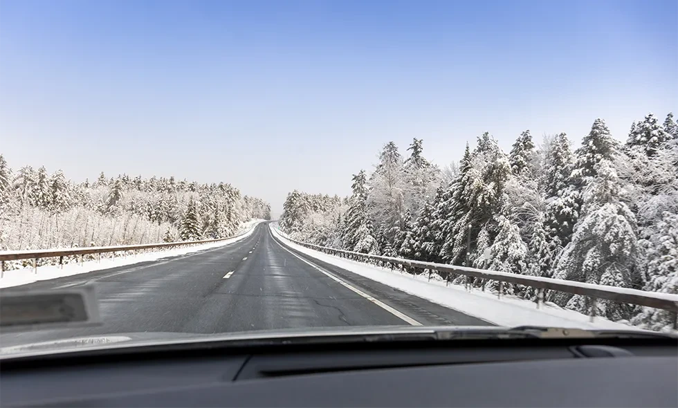 Rak landsväg med is, omgiven av snötäckta träd under klarblå himmel.
