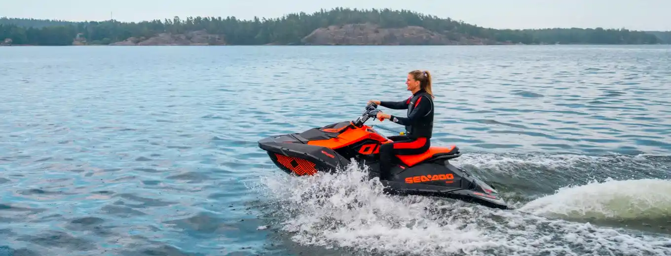 En kvinna kör en orange och svart vattenskoter på lugnt vatten, med skog och klippor i bakgrunden, klädd i en våtdräkt som skyddar mot stänk