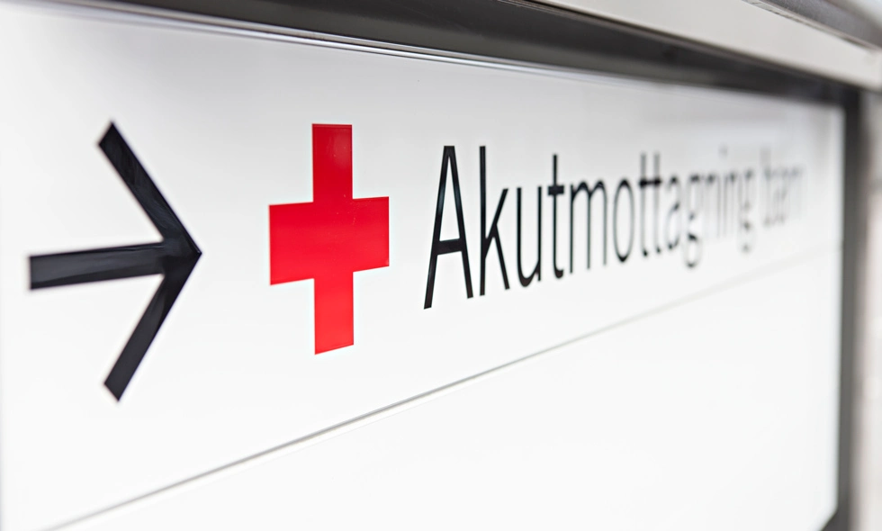 Skylt för akutmottagning med en röd kors-symbol och en pil som pekar åt höger, i en sjukhuskorridor