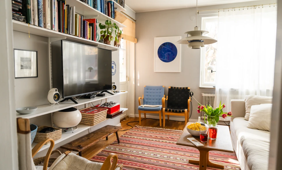 Ett vardagsrum med bokhylla, TV, blått konstverk, randig rag-matta och soffbord med tulpaner framför en vit soffa.