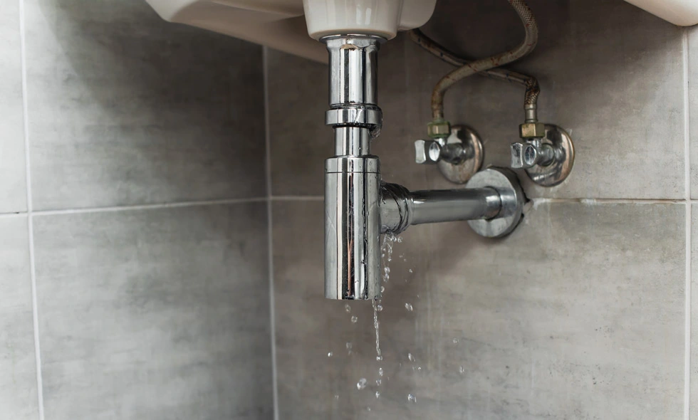 Vatten läcker från en kromad P-trap under ett handfat, med droppar som faller mot en grå kaklad golv, vilket indikerar behov av rörreparation eller underhåll